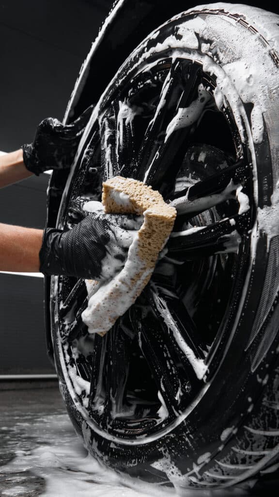 Lavage de Jante Noire : Nettoyage Mousseux avec Éponge Mains gantées nettoyant une jante noire de voiture avec une éponge savonneuse et mousse abondante.