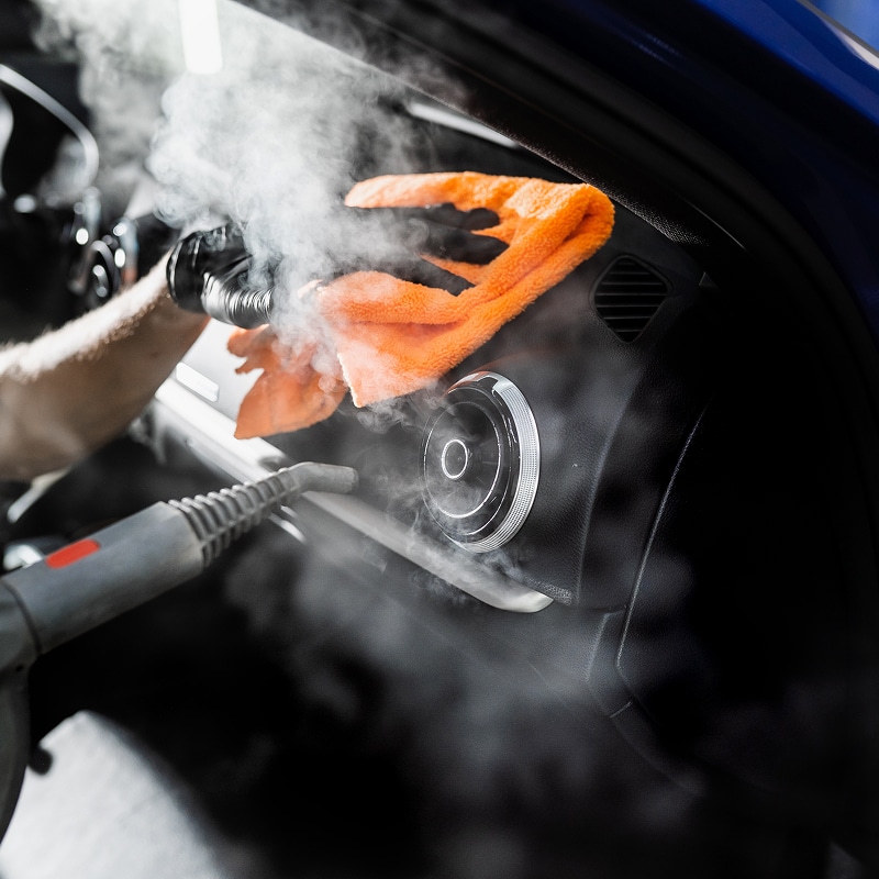 Nettoyage vapeur tableau de bord voiture : gant noir essuie une aération chromée avec un chiffon orange et un pistolet vapeur.