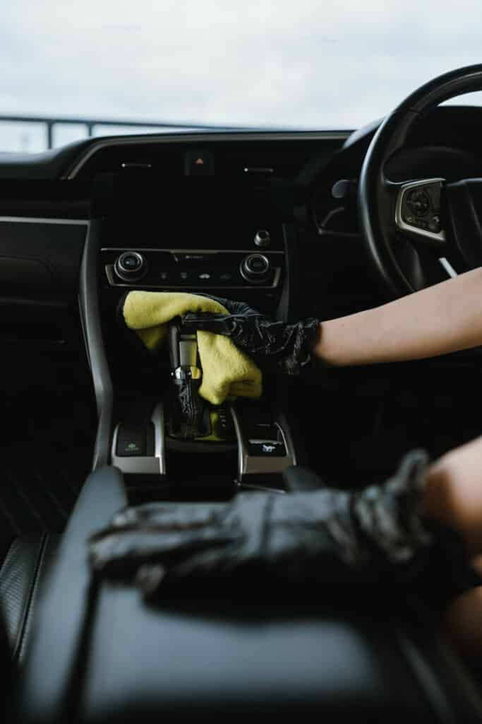 Nettoyage intérieur voiture : levier de vitesse et console Gant noir nettoyant le levier de vitesse (R, N, D, S, L) avec un chiffon microfibre jaune dans l'habitacle sombre d'une voiture.