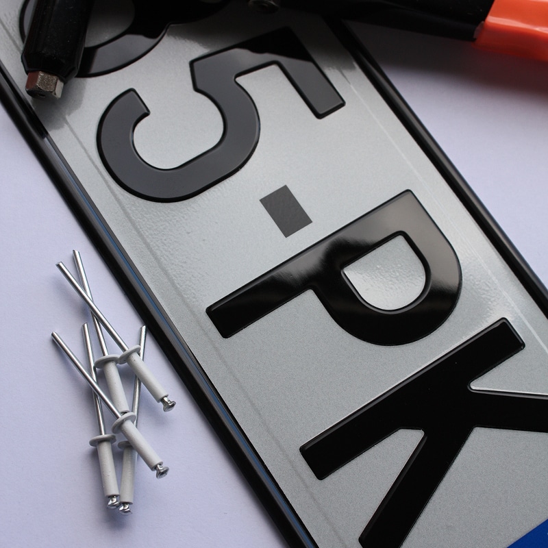 Plaque d'immatriculation style européen, gris argenté et caractères noirs en relief, avec rivets assortis à côté. Texte visible : 5-PK.