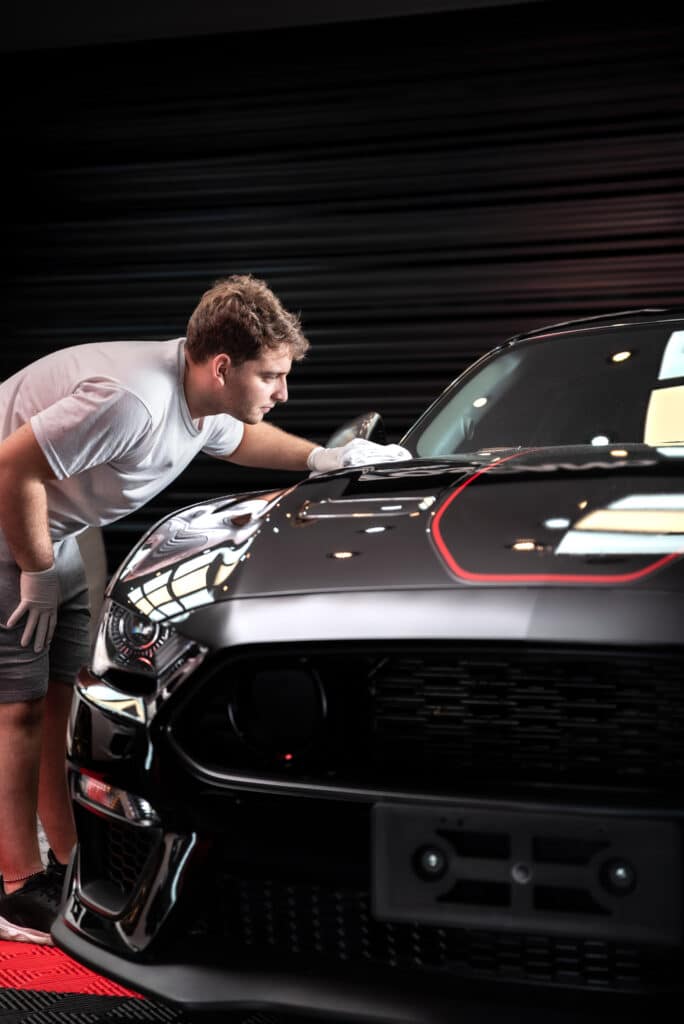 Polissage voiture noire : finition méticuleuse pro Jeune homme en t-shirt blanc et gants vérifie la finition d'une voiture de sport noire brillante dans un garage.